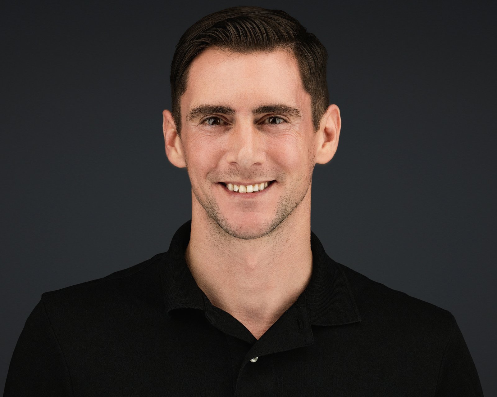 Smiling man in a black collared shirt in a professional headshot against a dark background