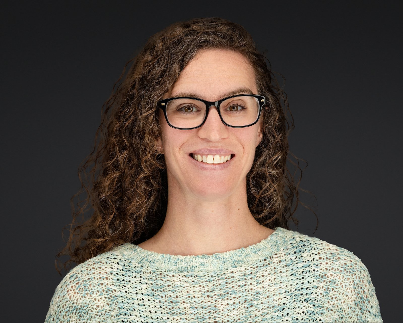 Woman with curly brown hair and glasses in a team headshot against a dark background